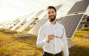 Um homem de camisa branca fica ao ar livre perto de painéis solares, segurando óculos.