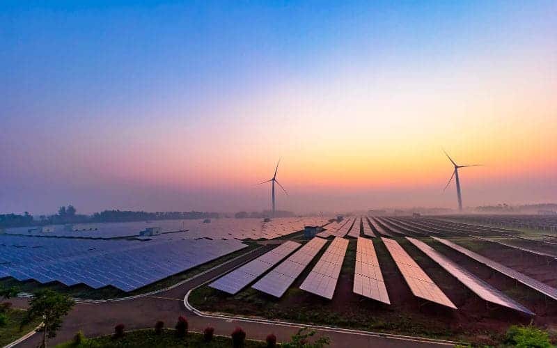 Ein Sonnenaufgang über einem Solarpark mit Windrädern im Hintergrund, der erneuerbare Energiequellen in einer ländlichen Landschaft präsentiert.