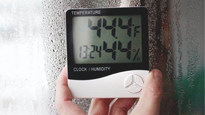 A person holds a digital hygrometer showing a temperature of 44.3°f and a humidity level of 38.4% against a foggy window.