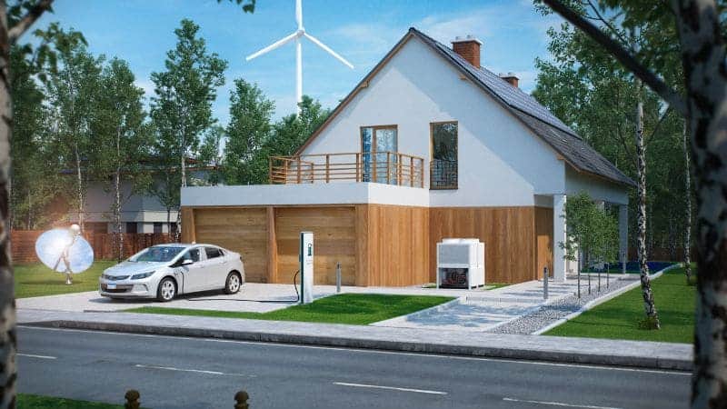 Casa moderna de dos plantas con garaje de madera, carga de coches eléctricos, paneles solares y un aerogenerador al fondo, situada en una zona suburbana.